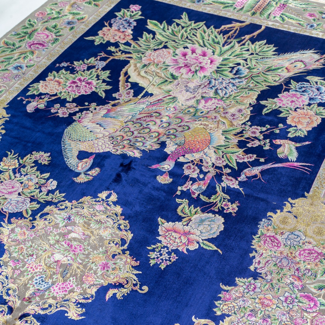 Luxurious silk rug with peacock pair and blooming florals on a navy base. Handwoven in soft hues with intricate botanical detailing.