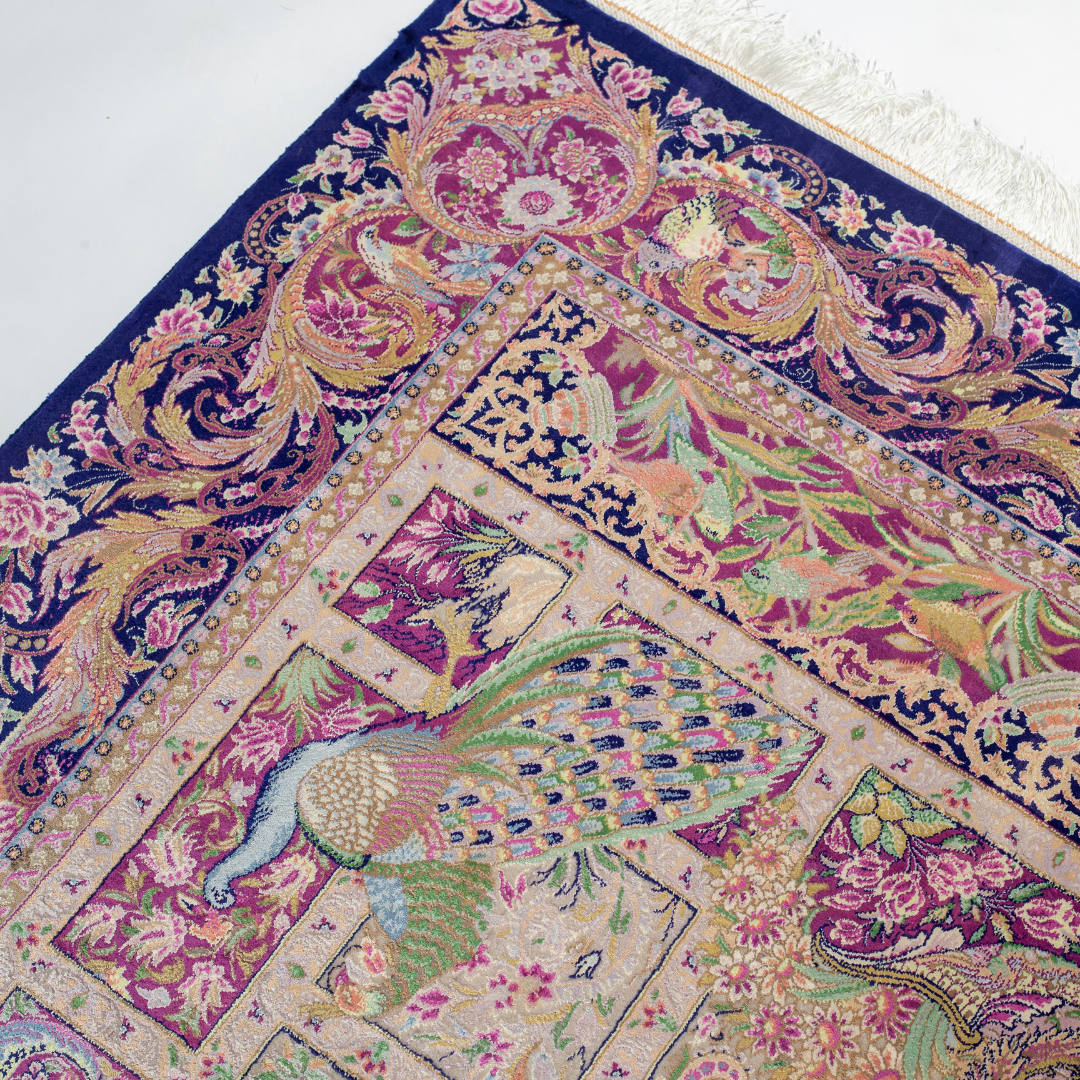 Luxurious silk rug with central bird tree, bordered by peacocks and florals. Handwoven in soft jewel tones with ornate framed details.