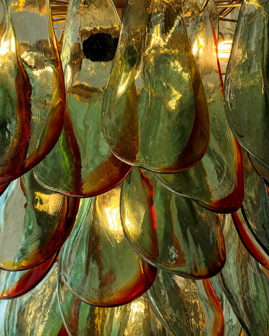 The Botanic Chandelier by The Qratist features handcrafted green and amber glass leaves layered around a refined metal frame, creating a sculptural botanical statement inspired by nature.