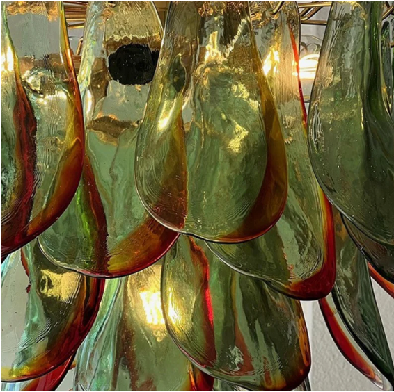 The Botanic Chandelier by The Qratist features handcrafted green and amber glass leaves layered around a refined metal frame, creating a sculptural botanical statement inspired by nature.
